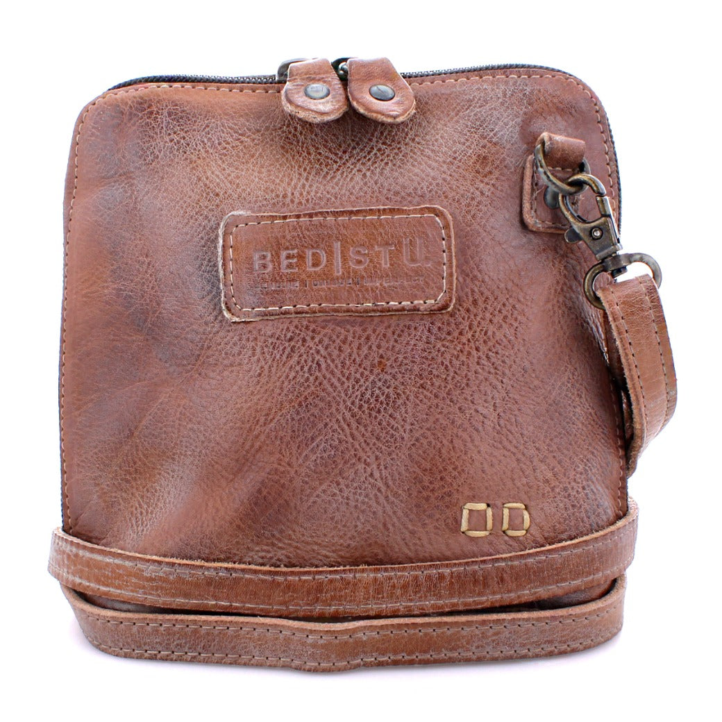 The Ventura crossbody bag by Bed|Stü is crafted from brown leather and features a zipper closure, a "Bed|Stü" logo patch, a metal clasp, and two small gold square accents at the bottom right corner. This lightweight purse also includes an exterior zip pocket for added convenience.
