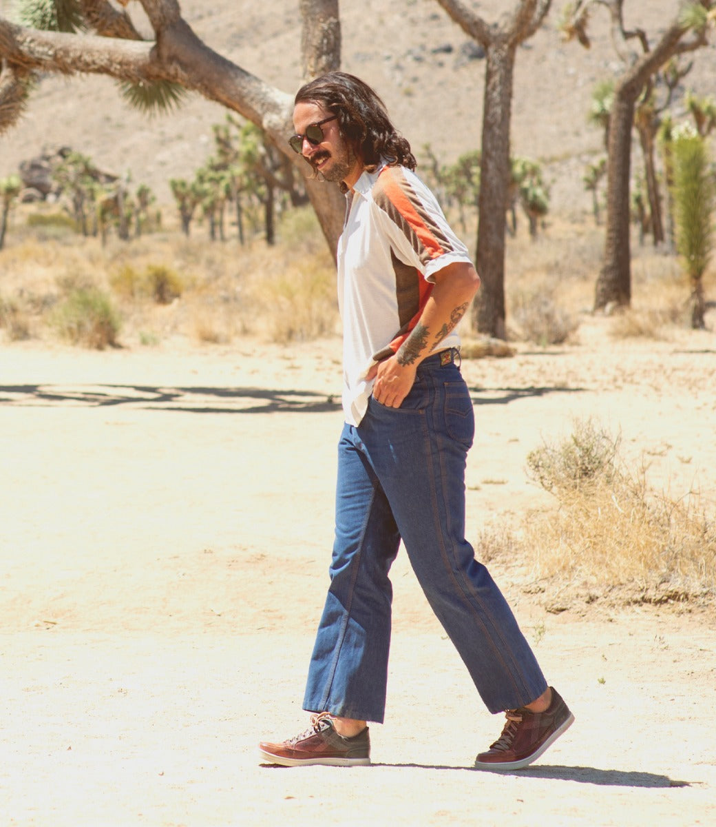 A man wearing sunglasses, a short-sleeved shirt, blue jeans, and Bed|Stü Wizard low-profile leather sneakers with crossover stitching walks in a dry, desert landscape with Joshua trees in the background.