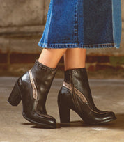 A person wearing the Bed|Stü Yuno boots—dark elevated heels with decorative stitching, a zipper, and contrast leather accents—paired with a blue denim skirt, standing on a concrete floor.