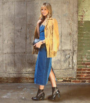 A woman stands indoors against a rustic brick and wood backdrop, wearing the Bed|Stü Yuno—elevated heel boots featuring contrast leather accents and breathable cushioning—paired with a tan fringe jacket, blue denim dress, and holding a black clutch.