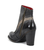 Introducing the Yuno by Bed|Stü—a black leather heeled ankle boot featuring a side zipper, decorative stitching, beige contrast leather accents, and a red interior lining, designed with breathable cushioning for comfort and shown from a back side angle.