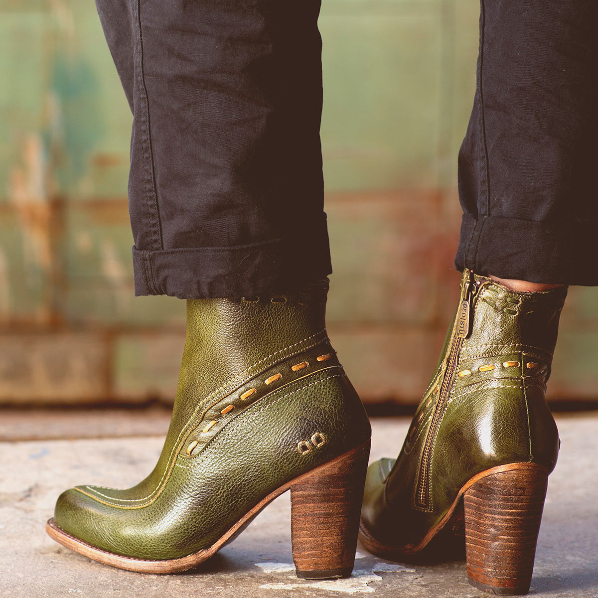 A person wearing the Bed|Stü Yuno green leather elevated heel boots featuring side zippers, contrast leather accents, and stitched details stands on a concrete surface.