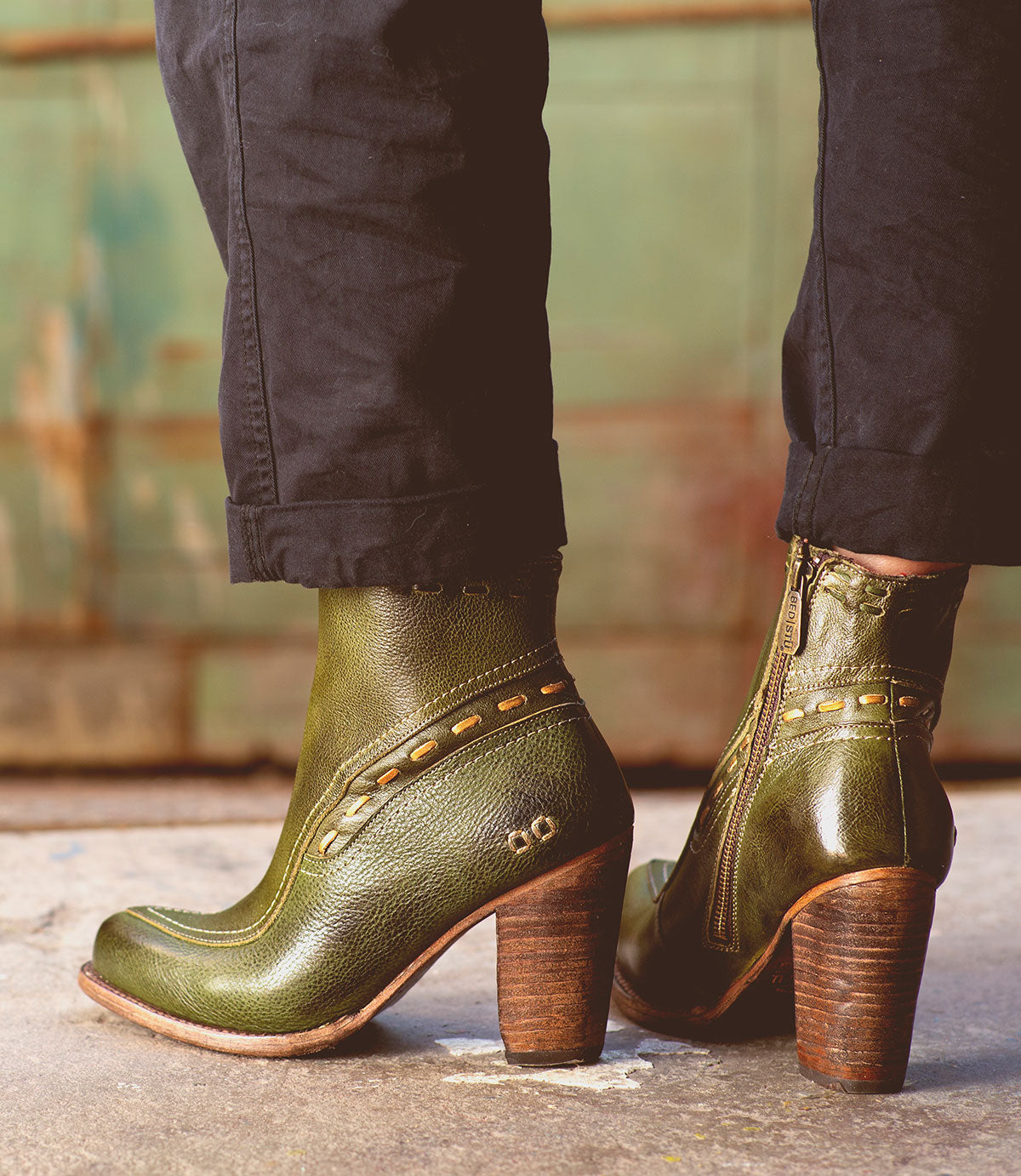 A person wearing the Bed|Stü Yuno green leather elevated heel boots featuring side zippers, contrast leather accents, and stitched details stands on a concrete surface.