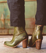 A person wearing the Bed|Stü Yuno green leather elevated heel boots featuring side zippers, contrast leather accents, and stitched details stands on a concrete surface.