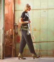 A woman in a black jumpsuit walks indoors, carrying the Yuno bag by Bed|Stü in green fringe and wearing matching green elevated heel boots with contrasting leather accents, set against a large distressed metal door.