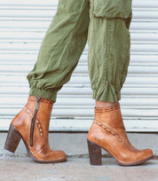 A person walks on a concrete surface in front of a white metal garage door, wearing olive green pants and the Bed|Stü Yuno tan elevated heel boots with side zippers and contrasting leather accents.