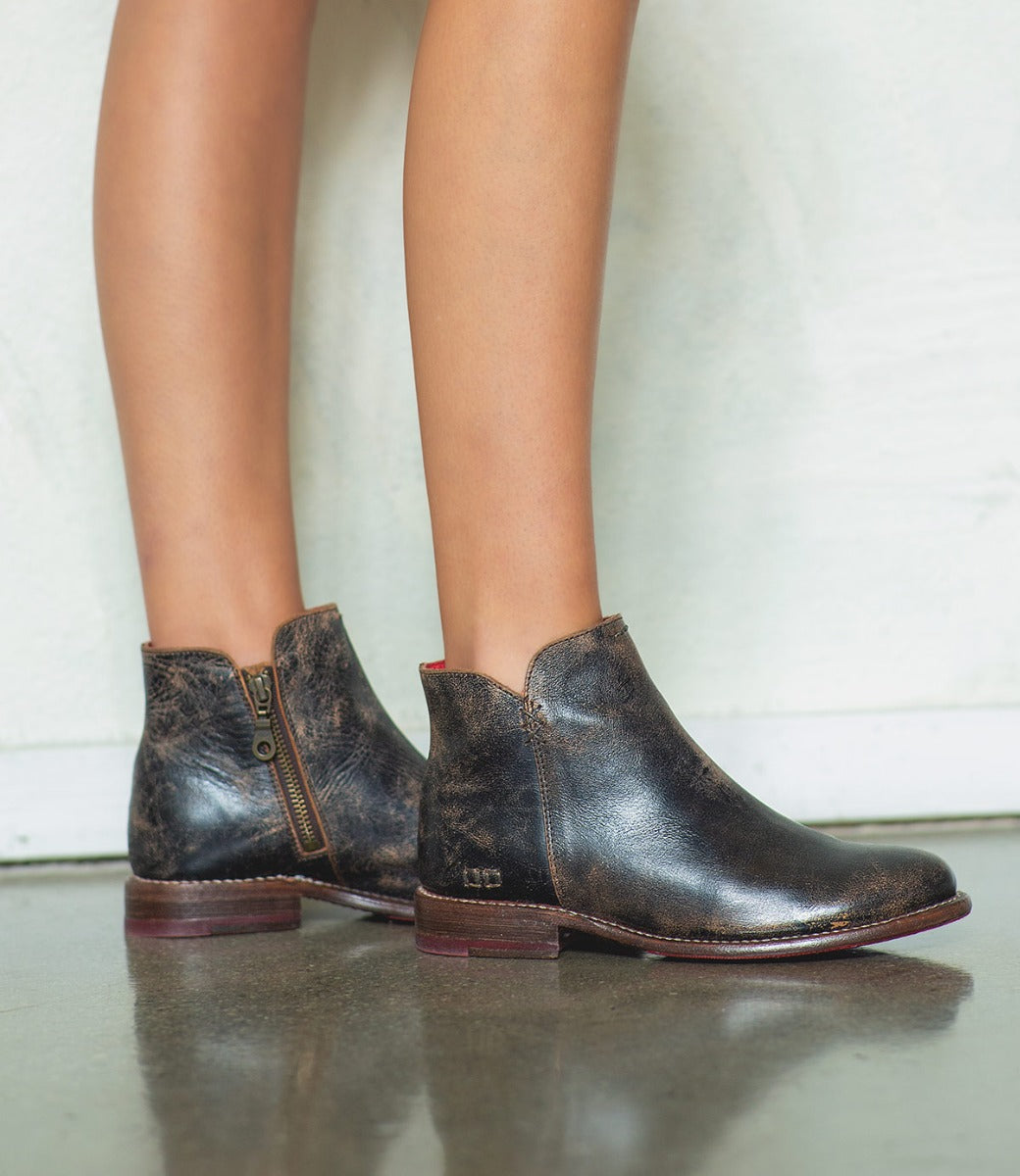 A person wearing the Yurisa handcrafted distressed black ankle boots by Bed|Stü, featuring side zippers and a durable leather outsole, stands on a shiny floor against a light-colored wall.