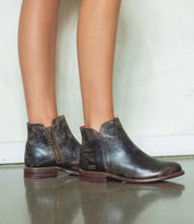 A person wearing the Yurisa handcrafted distressed black ankle boots by Bed|Stü, featuring side zippers and a durable leather outsole, stands on a shiny floor against a light-colored wall.