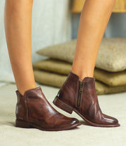 Wearing the handcrafted Yurisa ankle boot by Bed|Stü, featuring a leather outsole and side zippers, the person stands on a beige floor with tan cushions and a lamp in the background.