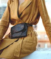 A person wearing a mustard-colored wrap dress accessorized with the Ziggy supple leather belt bag by Bed|Stü, seated outdoors with blurred buildings in the background.