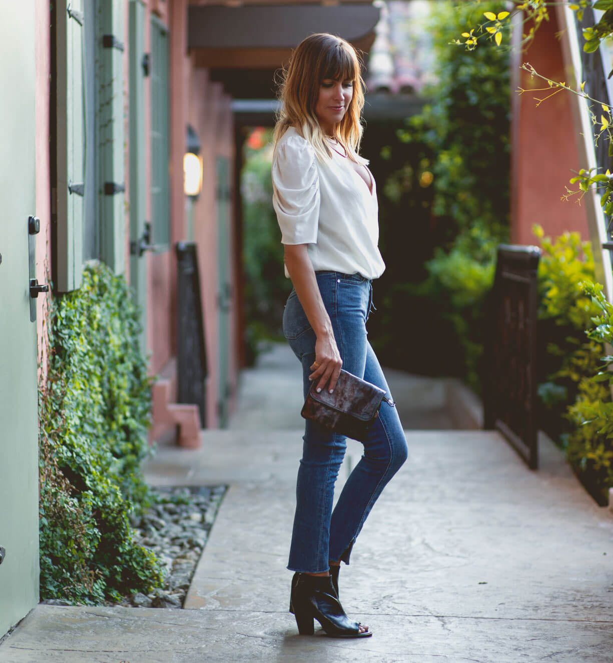 A woman stands sideways on a walkway outdoors, holding the chic Bed|Stü Ziggy crossbody bag. She wears a white blouse, blue jeans, and black open-toe boots, with greenery and buildings in the background.